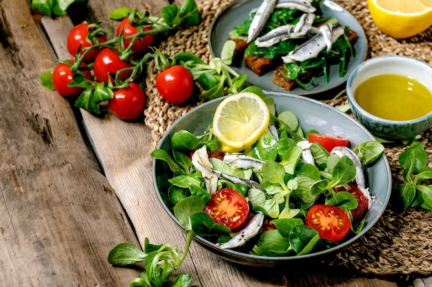 Foto ensalada de campo verde con filete de anchoas en escabeche o sardinas y tomates cherry, servido en un tazón azul con limón y aceite de oliva en una servilleta de paja sobre la mesa de madera vieja.