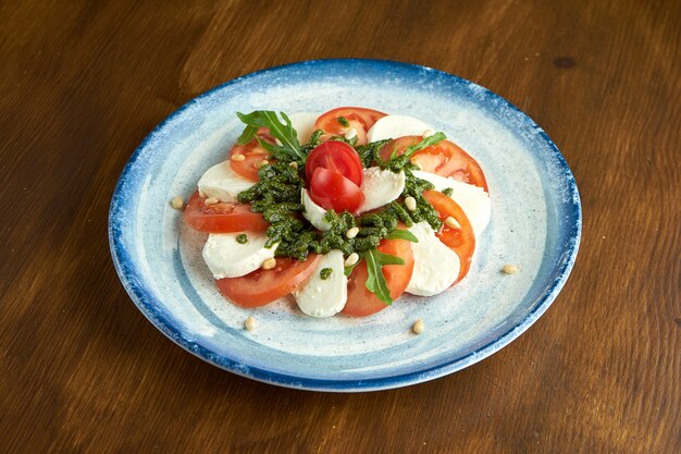 Ensalada caprese italiana clásica con tomate, mozzarella, rúcula y pesto en una placa azul sobre una superficie de madera