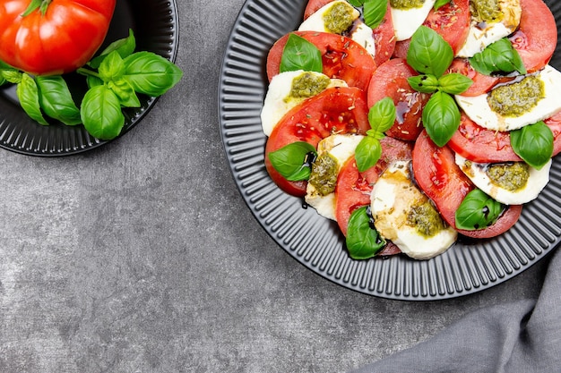 Ensalada caprese italiana tradicional con rodajas de tomate, mozzarella, albahaca, salsa pesto y especias sobre un fondo de hormigón gris oscuro. Vista superior. Copie el espacio.