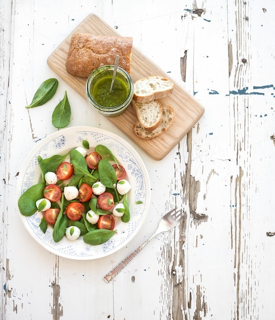 Ensalada caprese, rodajas de chiabatta, salsa de pesto casera, tomates cherry, espinacas y mozzarella en plato de cerámica sobre fondo rústico de madera blanca, vista superior