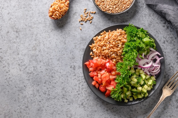 Ensalada de cereal integral deletreado con verduras frescas de verano, tomate, pepino, verduras en una mesa gris con espacio para texto. Vista desde arriba.