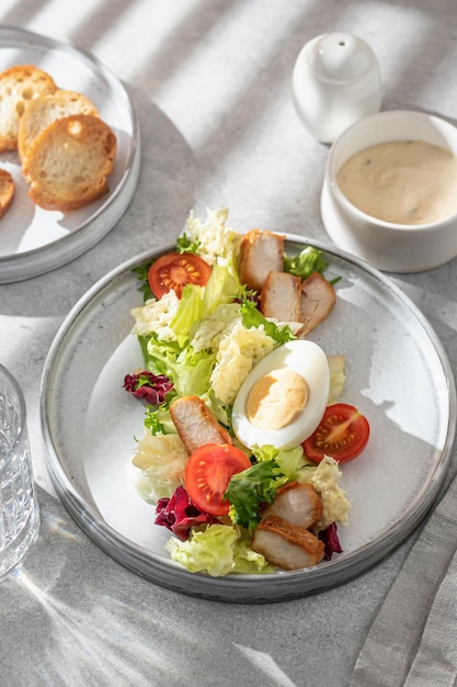 Ensalada César en alimentación lineal moderna Ensalada saludable con huevo de pollo, tomates, lechuga y chips de parmesano con cubiertos en una mesa gris a la luz de la mañana con sombra de ventana Menú de dieta ligera