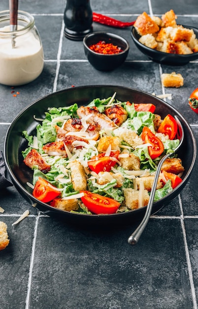 Ensalada César casera con pollo crujiente y tomates sobre un fondo de azulejos negros