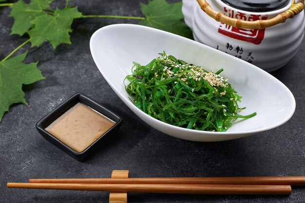 Ensalada de chuka con pepinos, semillas de sésamo y salsa, en un plato blanco, con una tetera japonesa, palillos y hojas de arce, sobre hormigón negro