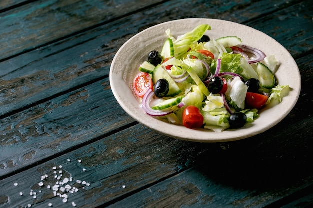 Ensalada clásica de verduras con tomate, pepino, cebolla, hojas de ensalada y aceitunas negras en plato de cerámica blanca. Mesa de madera oscura.