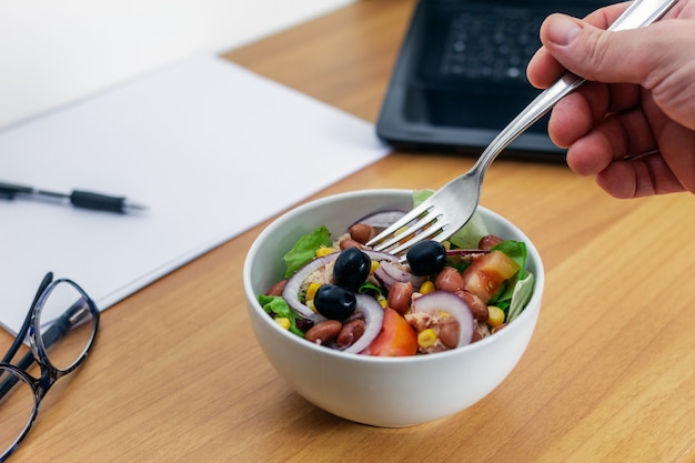 Una ensalada en un cuenco de porcelana blanca sobre un escritorio de oficina de madera con un empleado tomando un bocado. Al fondo, algunos útiles de oficina.