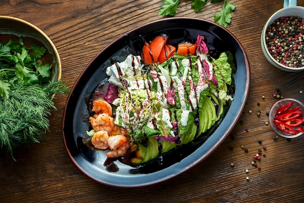 Ensalada deliciosa y dietética con camarones a la parrilla, queso feta, tomate y aguacate servido en un plato negro sobre una mesa de madera. Comida de restaurante