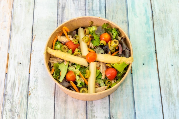 Ensalada con espárragos blancos, aceitunas picadas, tomates cherry, bonito del norte enlatado y brotes de lechuga variados con zanahorias en un recipiente de cartón para entrega a domicilio.