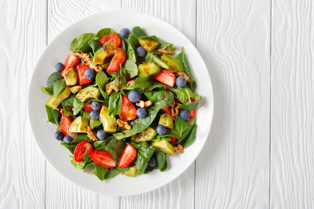 Ensalada de espinacas, aguacate, nueces y bayas con vinagre balsámico y aderezo de semillas de amapola en una placa blanca.