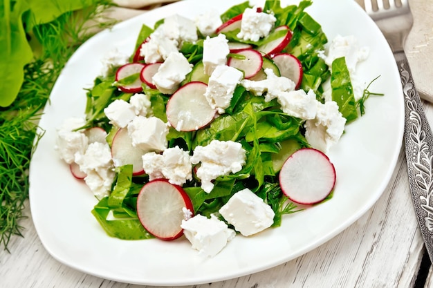 Ensalada con espinacas, pepinos, rábanos y queso salado, eneldo y cebollas verdes en un plato blanco, una toalla al fondo de una tabla de madera clara