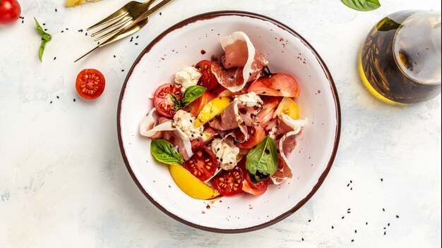 Ensalada fresca saludable de tomates vegetales, bolas de queso crema, jamón prosciutto y hojas de albahaca. Fondo de comida para menú, receta. Vista superior.