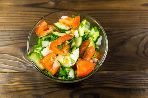 Ensalada fresca con tomate, pepino, cebolla, perejil y eneldo en un tazón de vidrio sobre mesa de madera