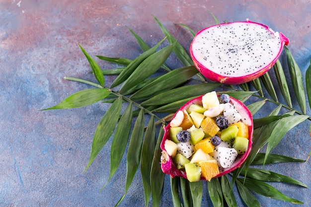 Ensalada de frutas exóticas servida en la mitad de una fruta de dragón en hojas de palma en el fondo de piedra, espacio de copia