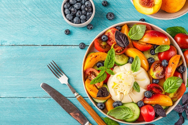 Ensalada de frutas de verano saludable con queso burrata tomates pepino y albahaca sobre mesa azul con cubiertos Vista superior