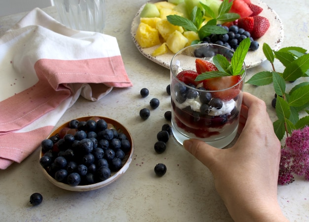 Ensalada de frutas y yogur griego natural con bayas frescas mano sosteniendo un vaso con yogur y frutas