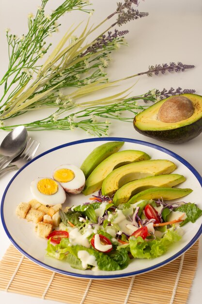 La ensalada grande consiste en rodajas de aguacate, huevo cocido, lechuga, lechuga, tomate y bizcochos, cubierta con crema de ensalada, aguacate cortado por la mitad en la espalda, Una comida con muchas verduras.