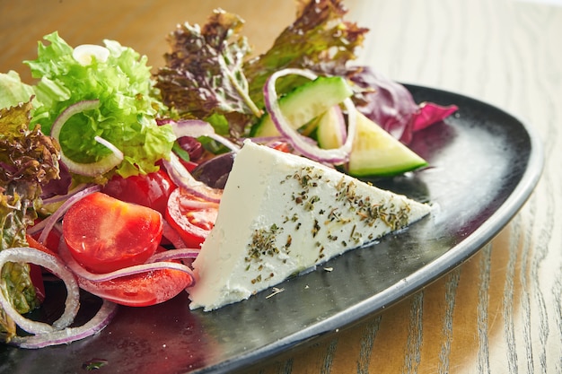 Una ensalada griega clásica y saludable con queso feta casero, cebolla roja, tomate y lechuga en un plato negro sobre una pared de madera. Primer plano, enfoque selectivo