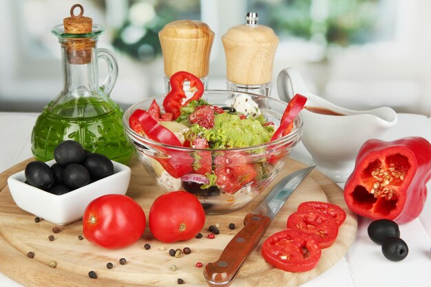 Foto ensalada griega fresca en un recipiente de vidrio rodeado de ingredientes para cocinar en una mesa de madera en el fondo de la ventana de cerca