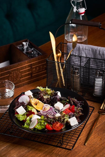 Ensalada griega fresca con tomate, rábano, cebolla, pimiento, aceitunas y queso feta en placa negra, vista lateral, fondo de madera