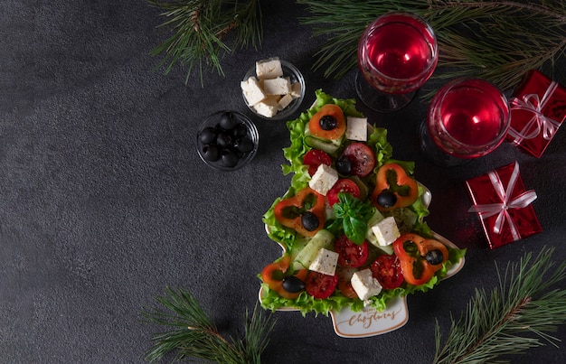 Ensalada griega saludable servida en plato como árbol de Navidad con decoración festiva y dos copas de vino sobre fondo oscuro. Copia espacio