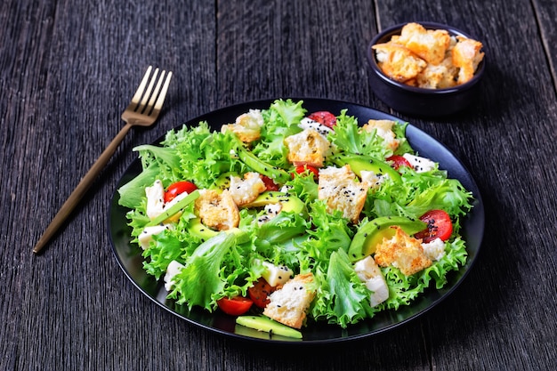 Ensalada de hojas con aguacate, tomates cherry, mozzarella y picatostes espolvoreados con semillas de sésamo negro en un plato, sobre una mesa de madera, vista horizontal desde arriba