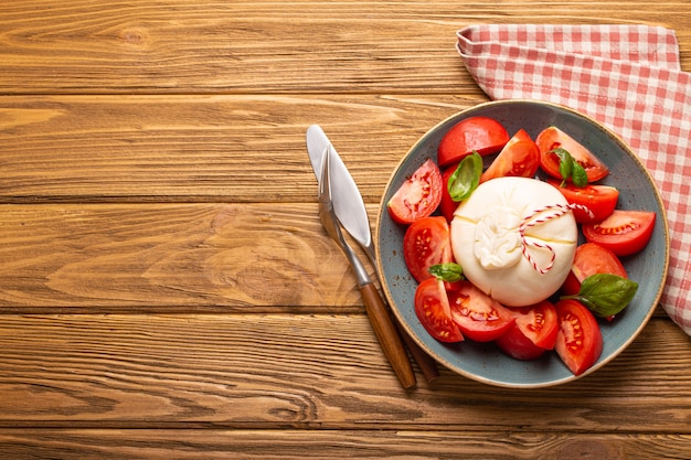 Ensalada italiana con tomates frescos, queso burrata, albahaca y aceite de oliva servido en un plato de cerámica sobre fondo de madera rústica desde arriba, refrigerio saludable o almuerzo con espacio para texto