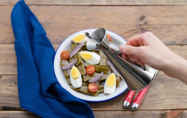 Ensalada de judías verdes con huevo cocido y tomate. Mano de mujer con aceitera. Copie el espacio. Fondo de madera.