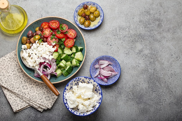 Ensalada mediterránea griega con tomates, queso feta, pepino, aceitunas enteras y cebolla roja en placa de cerámica azul sobre fondo de hormigón gris desde arriba, aperitivo tradicional de Grecia con espacio de copia