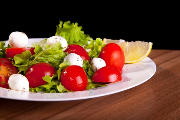 Ensalada mediterránea de verduras con bolas de queso y limón, tomates, verduras, sobre una mesa de madera y fondo negro.