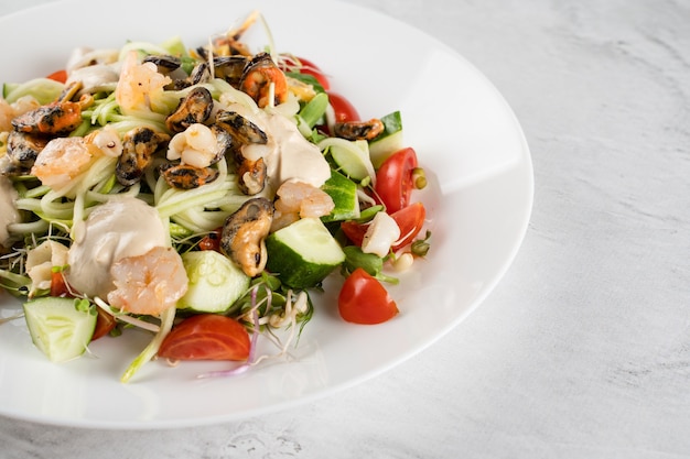 Ensalada con mejillones, camarones, tomate cherry, pepino, pasta de calabacín, microgreen, salsa de yogur en plato blanco sobre mesa de luz.