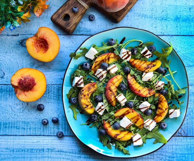 Ensalada de melocotón a la parrilla con queso feta, arándanos, rúcula y aceite de oliva sobre un fondo azul.