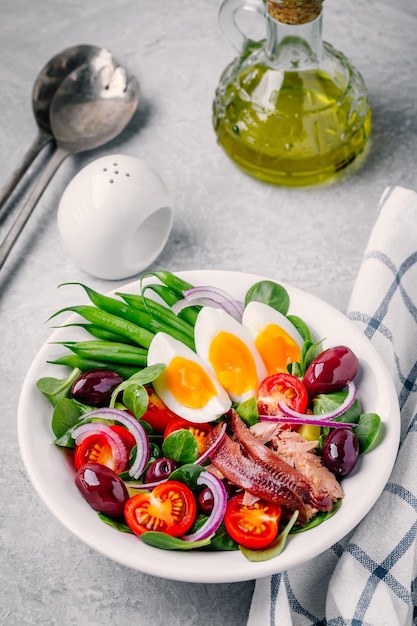 Ensalada Nicoise con atún, anchoas, huevos, judías verdes, aceitunas, tomates, cebollas rojas y hojas de ensalada sobre fondo gris.