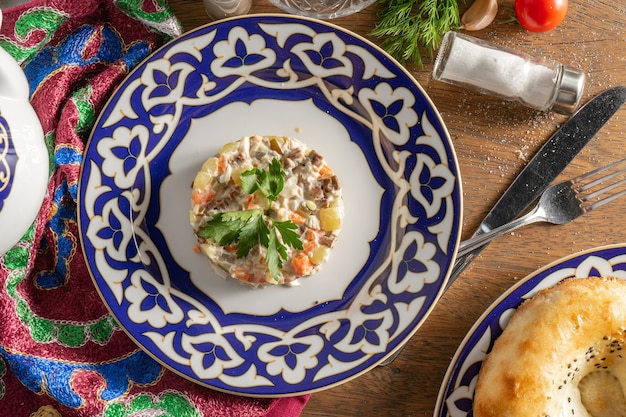 Foto ensalada olivier clásica con mayonesa, verduras y salchicha decorada con perejil en un plato de cerámica con adornos tradicionales uzbecos sobre una mesa de madera.