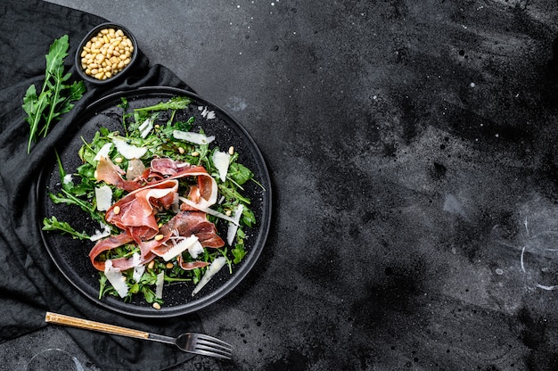 Ensalada de Parma, jamón, rúcula y parmesano. Fondo negro, vista superior, espacio para texto