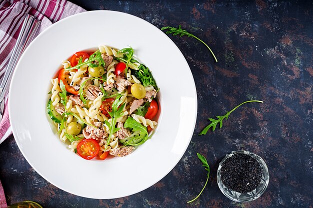 Ensalada de pasta con atún, tomate, aceitunas, pepino, pimiento y rúcula sobre fondo rústico. Vista superior