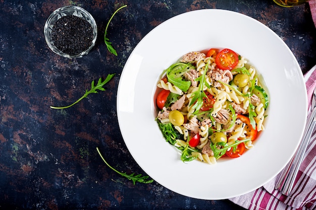 Ensalada de pasta con atún, tomates, aceitunas, pepino, pimiento dulce y rúcula sobre fondo rústico