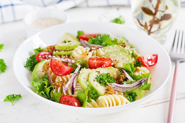 Ensalada de pasta fusilli con tomates aguacate lechuga verde fresca cebolla roja y aderezo de mostaza sobre fondo blanco Almuerzo vegetariano saludable