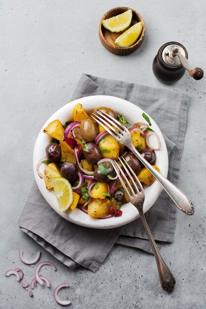 Ensalada de patata caliente con aceitunas, pimiento, perejil y cebolla roja sobre placa de cerámica blanca antigua sobre fondo de hormigón gris