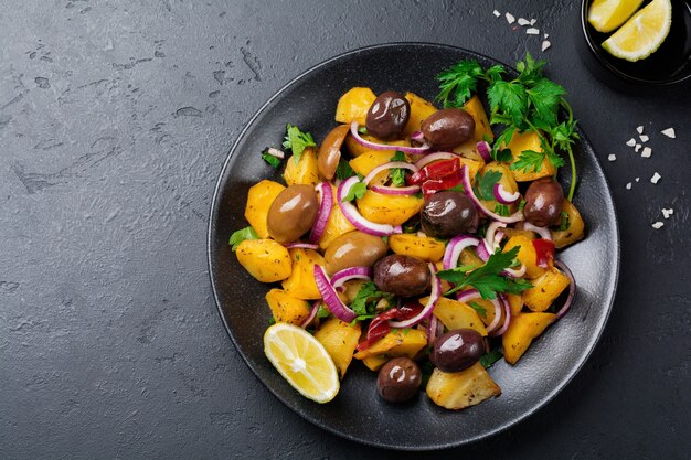 Ensalada de patata caliente con aceitunas, pimiento, perejil y cebolla roja sobre placa de cerámica negra sobre superficie de hormigón oscuro