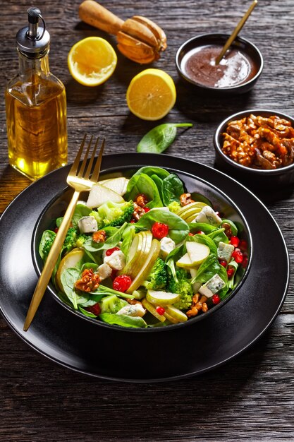 Ensalada de pera con cubos de queso azul, brócoli, hojas de espinaca, nueces caramelizadas y semillas de granada en un plato negro sobre la mesa de madera, guarnición de acción de gracias, vista vertical desde arriba