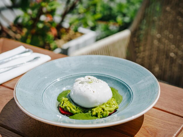 Foto ensalada de queso buratta con base de crema de aguacate en plato rústico azul en restaurante o cafetería burratina comida fresca y saludable dieta de merienda en mesa de madera en verano día soleado espacio para copiar al aire libre