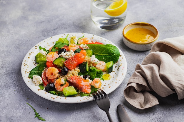 Ensalada con quinua, lechuga iceberg, rúcula, pepino, aceitunas negras, tomate, requesón, salmón, camarones y salsa de mango en la pared gris. alimentación limpia para aumentar la inmunidad