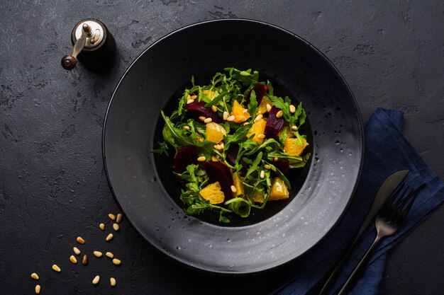 Ensalada de remolacha, naranja, piñones, aceite de oliva, queso feta y rúcula en placa de cerámica negra sobre fondo de mesa de hormigón oscuro. Vista superior.