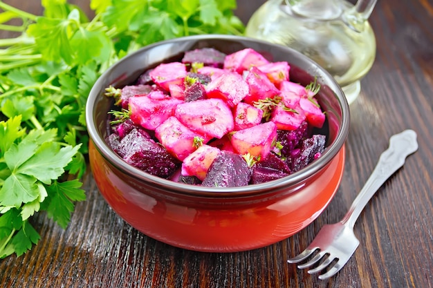 Foto ensalada de remolacha y papa, sazonada con aceite vegetal y vinagre en un bol, perejil, tenedor contra una tabla de madera oscura.