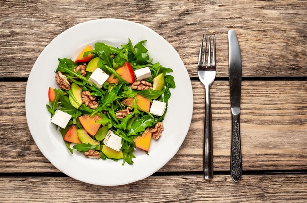 Ensalada de rúcula, caqui, nueces, aguacates, queso feta en un plato blanco sobre una mesa de madera