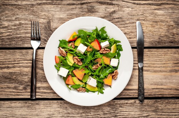 Ensalada de rúcula, caqui, nueces, aguacates, queso feta en un plato blanco sobre una mesa de madera