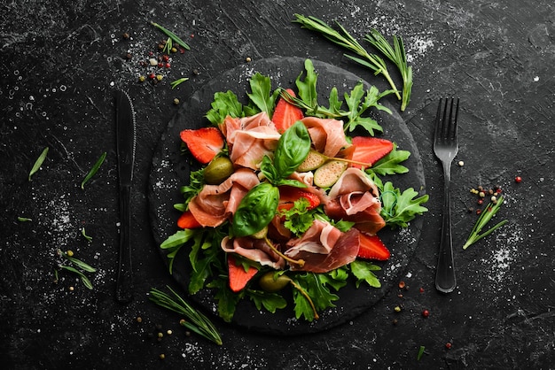 Ensalada de rúcula prosciutto fresas y alcaparras en un plato de piedra negra cocina italiana