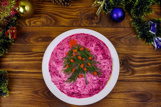 Ensalada rusa tradicional Arenque bajo un abrigo de piel shuba y adornos navideños en una mesa de madera Vista superior Ensalada decorada con árbol de Navidad hecho de eneldo y zanahoria