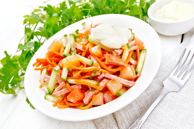 Ensalada de salchicha ahumada, zanahoria picante, tomate, pepino y especias con mayonesa, servilleta, tenedor y perejil sobre un fondo de tabla de madera