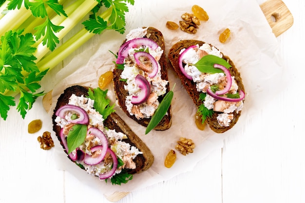 Ensalada de salmón, peciolo de apio, pasas, nueces, cebollas rojas y requesón sobre pan tostado con lechuga verde sobre papel sobre una tabla de madera de fondo desde arriba